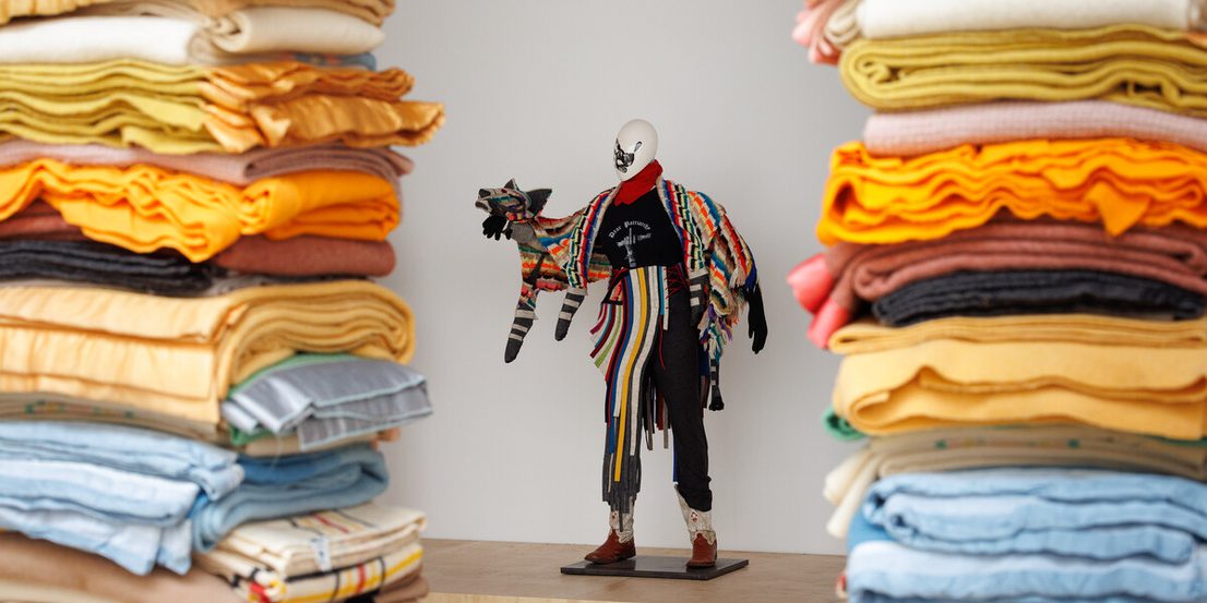 A mannequin dressed in a colourful, eclectic outfit stands on a wooden platform. It holds a striped, stuffed animal. Piles of folded, multicoloured fabrics frame the scene on both sides, creating a vibrant and artistic display.