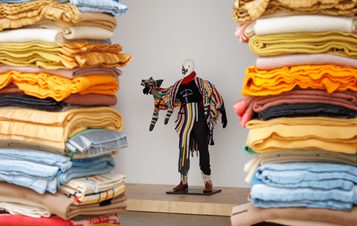 A mannequin dressed in a colourful, eclectic outfit stands on a wooden platform. It holds a striped, stuffed animal. Piles of folded, multicoloured fabrics frame the scene on both sides, creating a vibrant and artistic display.