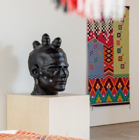A gallery displays a black sculpted head with geometric patterns on a pedestal. In the background, a colourful textile with intricate designs hangs on the wall. Foreground shows part of another textile with red, orange, and white patterns.