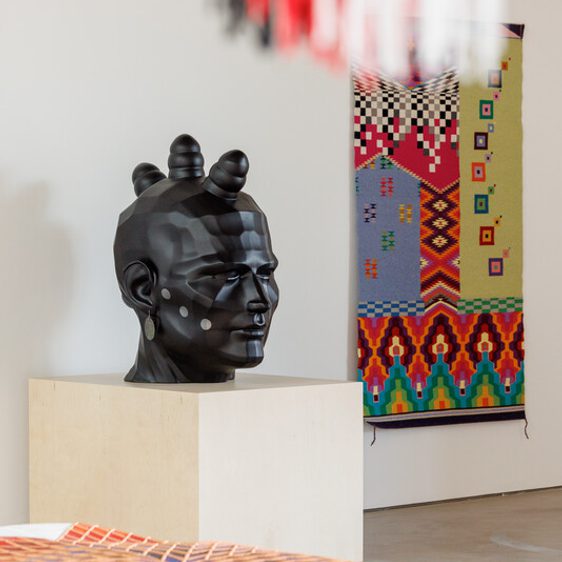 A gallery displays a black sculpted head with geometric patterns on a pedestal. In the background, a colourful textile with intricate designs hangs on the wall. Foreground shows part of another textile with red, orange, and white patterns.