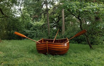 A wooden rowboat with oars is placed on a grassy area surrounded by lush green trees. The boat appears well-crafted and polished, contrasting with the natural forest setting.