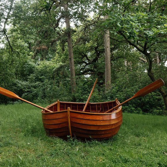 A wooden rowboat with oars is placed on a grassy area surrounded by lush green trees. The boat appears well-crafted and polished, contrasting with the natural forest setting.