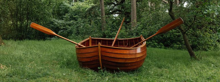A wooden rowboat with oars is placed on a grassy area surrounded by lush green trees. The boat appears well-crafted and polished, contrasting with the natural forest setting.