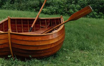 A wooden rowing boat with oars rests on lush green grass, surrounded by dense trees and foliage. The scene is tranquil, with the boat's rich brown hue contrasting against the vibrant greenery of the forest.