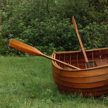 A wooden rowboat with two oars rests on lush green grass, surrounded by dense trees and foliage. The boat's rich brown hue contrasts with the vibrant greenery, creating a serene, natural setting.