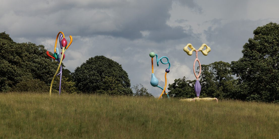 A large, abstract sculpture featuring vibrant, curved shapes in various colours, resembling flowers or plant forms. It stands on a grassy area with trees in the background under a cloudy sky, creating a striking visual contrast.