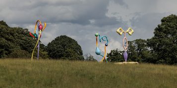 A large, abstract sculpture featuring vibrant, curved shapes in various colours, resembling flowers or plant forms. It stands on a grassy area with trees in the background under a cloudy sky, creating a striking visual contrast.