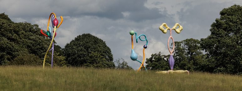 A large, abstract sculpture featuring vibrant, curved shapes in various colours, resembling flowers or plant forms. It stands on a grassy area with trees in the background under a cloudy sky, creating a striking visual contrast.