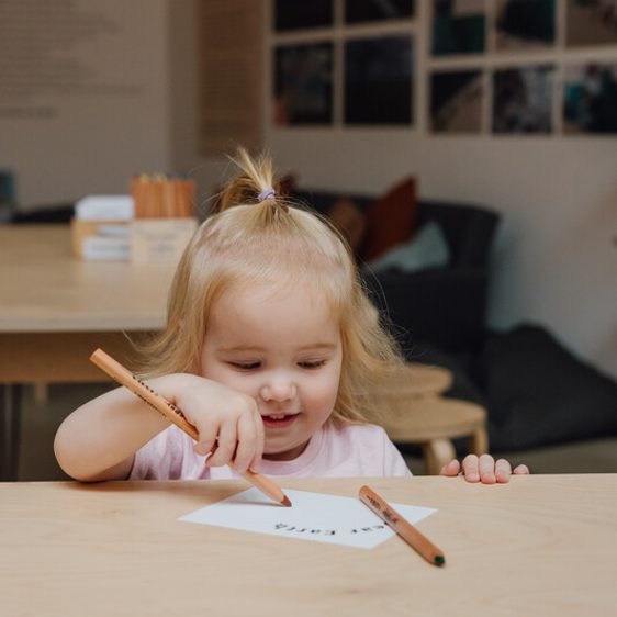 A young child with blonde hair sits at a table, holding a coloured pencil and drawing on a piece of paper. The room has a cosy atmosphere with blurred images on the wall and a sofa in the background.