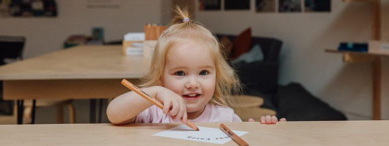 A young girl with blonde hair sits at a wooden table, smiling while drawing with coloured pencils on a piece of paper. The room has a cosy atmosphere with a sofa and photos on the wall in the background.