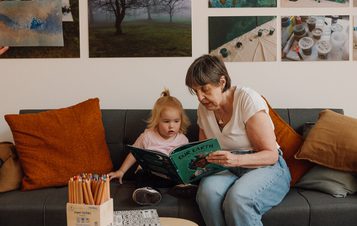 A woman and a young child sit on a grey sofa, reading a book titled "Our Earth". The room has orange cushions and a wall adorned with nature-themed photos. A box of coloured pencils is on the table in front of them.
