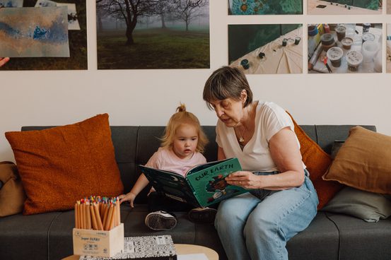 A woman and a young child sit on a grey sofa, reading a book titled "Our Earth". The room has orange cushions and a wall adorned with nature-themed photos. A box of coloured pencils is on the table in front of them.