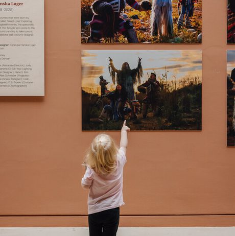 A young child with blonde hair points at a wall displaying two large photographs of people in traditional costumes. The background is a warm, earthy tone, and a descriptive plaque is visible on the left side.