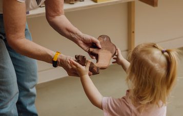 An adult hands a wooden toy to a young child with blonde hair in a ponytail. The child reaches out eagerly. They are indoors, with a wooden shelf in the background. The adult wears a yellow wristband and jeans.