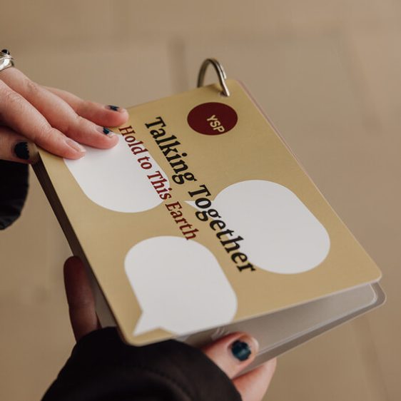 A person holds a beige booklet titled "Talking Together: Hold to This Earth" with speech bubble graphics. The booklet is bound with a metal ring. The person has dark nail polish and wears a dark jacket. The background is a light-coloured floor.