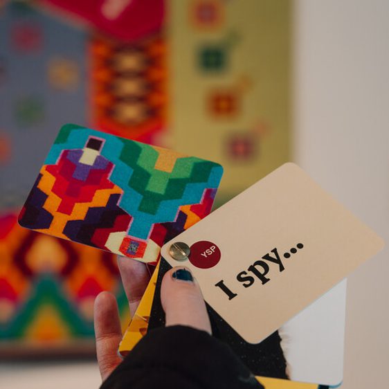 A hand holds colourful cards, one with a geometric pattern and another reading "I spy...". A blurred, vibrant tapestry is in the background. The person has dark nail polish and wears a dark sleeve.