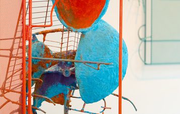 A colourful abstract sculpture featuring stacked, painted spheres and wire elements, presented within a red metal frame. A green sculpture is visible in the background.