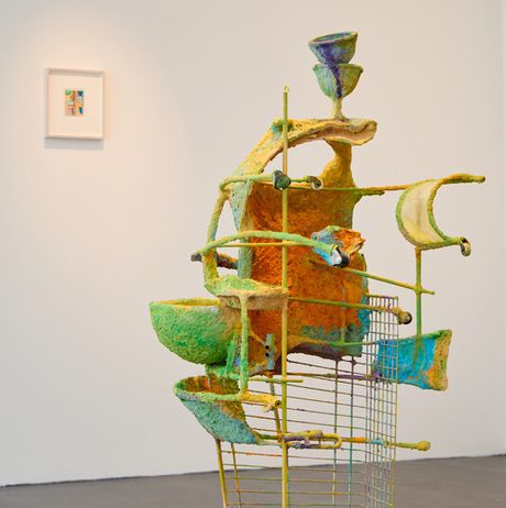 A colourful, abstract sculpture stands on a wheeled trolley within a gallery setting. The piece features layered, organic forms in shades of green, yellow, and blue.