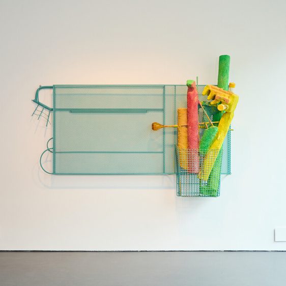 A turquoise metal frame holds a collection of brightly coloured, tubular objects – orange, red, green, and pink – within a wire basket. The artwork is displayed on a plain white wall.