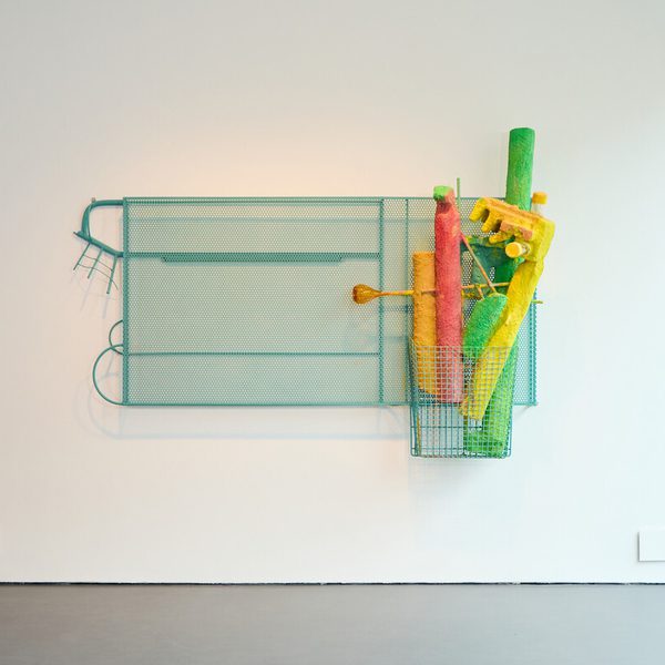 A turquoise metal frame holds a collection of brightly coloured, tubular objects – orange, red, green, and pink – within a wire basket. The artwork is displayed on a plain white wall.