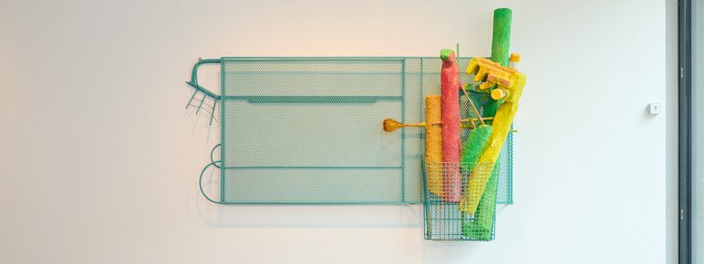 A turquoise metal frame holds a collection of brightly coloured, tubular objects – orange, red, green, and pink – within a wire basket. The artwork is displayed on a plain white wall.