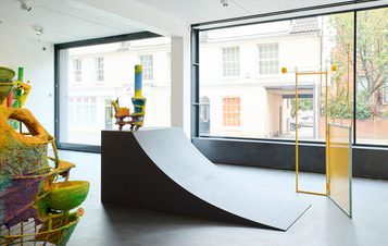 Contemporary art installation within a gallery space. A sculptural ramp and a yellow mesh structure are displayed alongside a colourful, textured sculpture. A large window provides a view of a street scene.