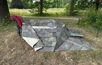 A weathered, sculptural installation featuring a section of slate roof and a red armchair, set in a grassy parkland. Trees are visible in the background.