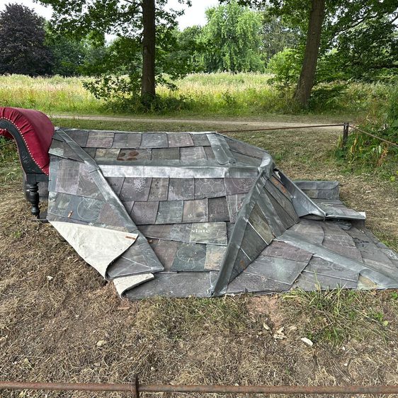 A weathered, sculptural installation featuring a section of slate roof and a red armchair, set in a grassy parkland. Trees are visible in the background.