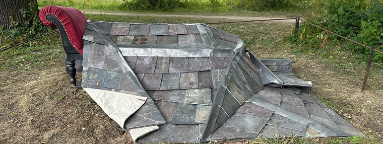 A weathered, sculptural installation featuring a section of slate roof and a red armchair, set in a grassy parkland. Trees are visible in the background.