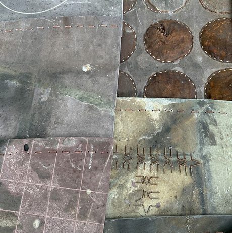 Close-up shot of four weathered metal plates. Each plate features circular cutouts and etched markings, suggesting a decorative or possibly industrial design.