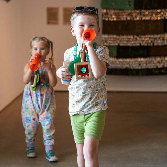 Two young children, a boy and a girl, are enjoying themselves in a gallery setting. The boy is blowing into an orange trumpet, whilst holding a wooden badge.