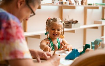 A young girl with pigtails is intently focused on a craft activity, with an adult assisting her. The scene is a bright, light-filled room.