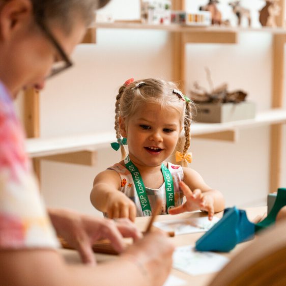 A young girl with pigtails is intently focused on a craft activity, with an adult assisting her. The scene is a bright, light-filled room.