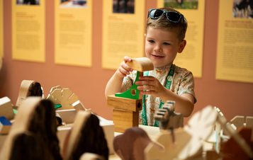 A young boy wearing sunglasses is intently crafting a wooden model with tools, surrounded by similar creations. The background is a muted wall with artwork.