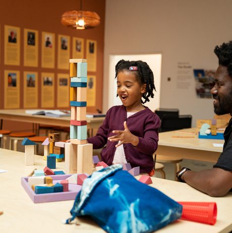 A young girl with braided hair is building a tower of blocks with her father in a bright, colourful room. Other children are visible in the background.