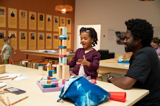 A young girl with braided hair is building a tower of blocks with her father in a bright, colourful room. Other children are visible in the background.