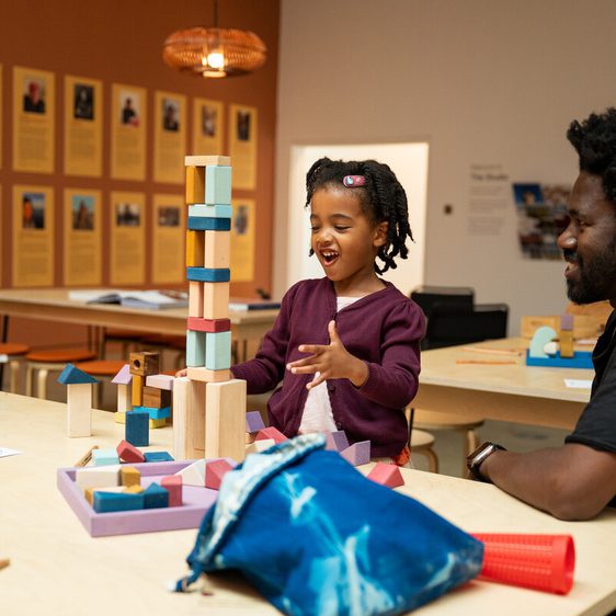 A young girl with braided hair is building a tower of blocks with her father in a bright, colourful room. Other children are visible in the background.
