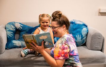 A woman reads a book to a young girl with pigtails, both smiling, on a grey sofa with blue cushions.