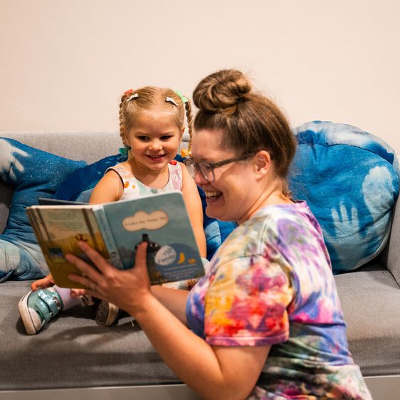A woman reads a book to a young girl with pigtails, both smiling, on a grey sofa with blue cushions.