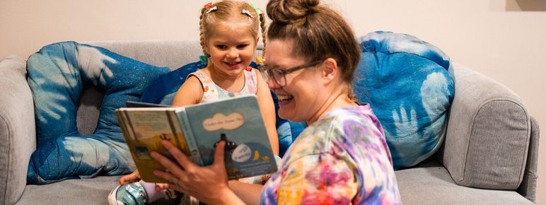 A woman reads a book to a young girl with pigtails, both smiling, on a grey sofa with blue cushions.