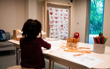 A young girl is seated at a table, drawing with coloured pencils. A quilt featuring red images is displayed on the wall behind her.