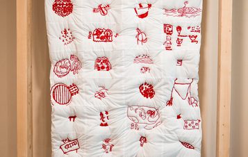 A white quilt with red embroidered images of various objects is displayed within a light-wood frame on wheels. The design features a repeating pattern.