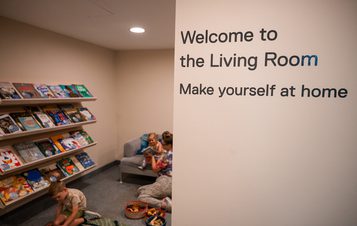 A welcoming living room scene with children reading on the floor, surrounded by bookshelves. A sign reads "Welcome to the Living Room Make yourself at home.