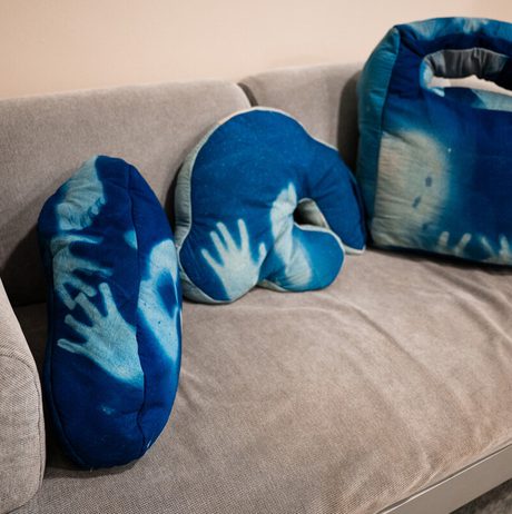 Three cushions in varying sizes are arranged on a grey sofa. Each cushion has a blue tie-dye pattern depicting a hand.