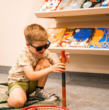 A young boy wearing sunglasses is playing with wooden blocks in a library setting, surrounded by bookshelves filled with children’s books.