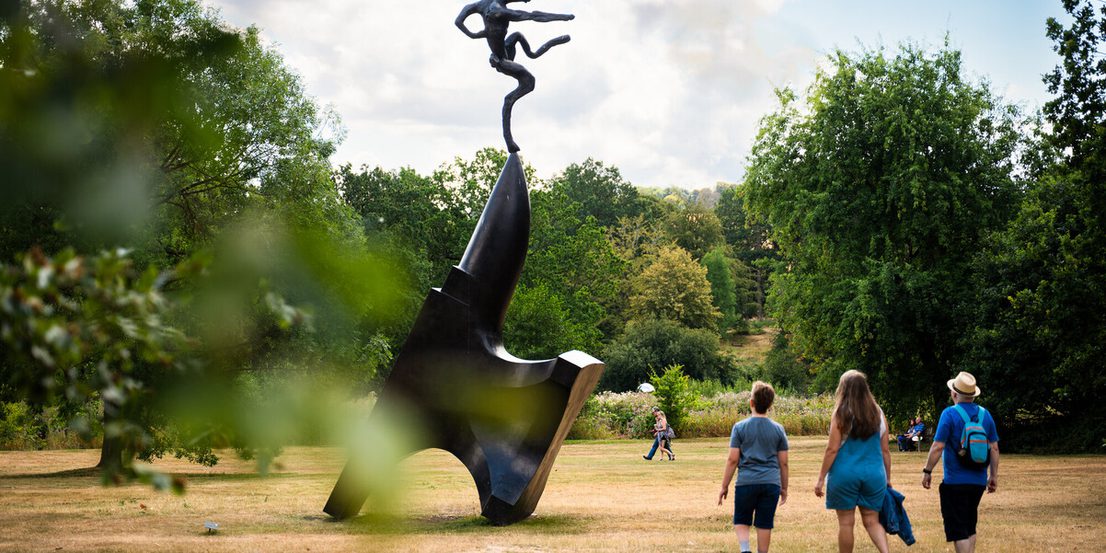 A group of three individuals walk away from a large, abstract sculpture in a park setting, with trees and a cloudy sky in the background.