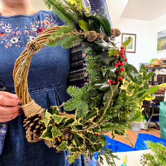 A woman holds a freshly made wreath of pine, fir, holly and pinecones. The wreath is in a woven basket and sits in a workshop setting.