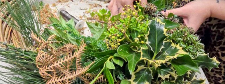 A person is arranging foliage – including holly, pine and ferns – onto a woven wreath. The scene is a workshop setting.