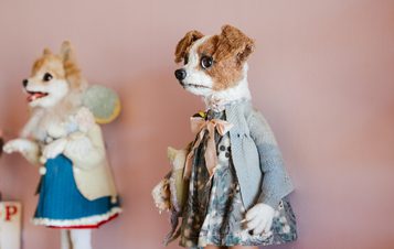 A close-up shot of a handmade dog puppet, dressed in a blue coat and patterned dress, standing against a pink wall. A painted dog figure is visible in the background.