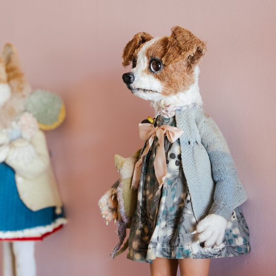 A close-up shot of a handmade dog puppet, dressed in a blue coat and patterned dress, standing against a pink wall. A painted dog figure is visible in the background.